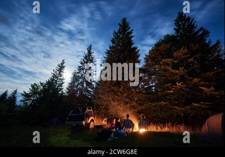 Group of friends near campfire, pickup offroad truck, tents at night camp in the mountains surrounded by spruce trees under magical starry sky, rear view. Car travel and camping concept Stock Photo