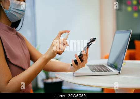 Asian businesswoman wearing protective Covid face covering spraying mobile phone screen with sanitizer, protection, health, hygiene Stock Photo