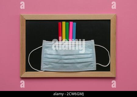 Face mask on top of colored pens and blackboard. Pink background. Stock Photo