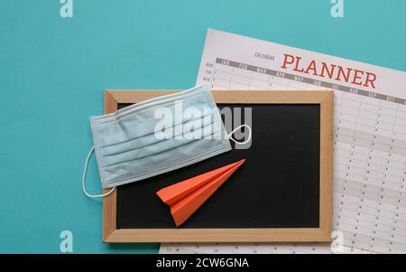 Face mask on top of blackboard and calendar planner with paper plane. Blue background. Stock Photo