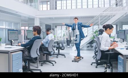 Young Happy Handsome Manager Wearing a Suit and Tie Rides a Longboard. Diverse and Motivated Business People Work on Computers in Modern Open Office. Stock Photo
