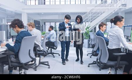 Couple of Young Male and Female Specialiasts Walk in the Office Holding a Tablet Computer, Ignore Their Colleague and Discuss Work. Motivated Diverse Stock Photo