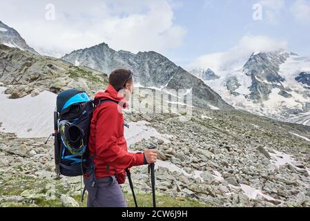 Side view snapshot of a man wearing a red jacket and a backpack, having a walk with hiking poles in the mountains situated in Swiss Alps, enjoying beautiful scenery