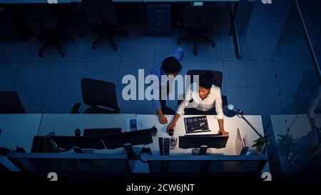 Working Late at Night in the Office: Businesswoman and Businessman Using Desktop Computer, Having Discussion, Analyzing, Using Documents, Solving Stock Photo