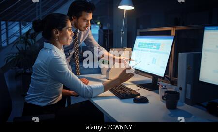 Late at Night In Modern Office: Businessman and Businesswoman Work on Desktop Computer, Having Discussion, Finding Problem Solution, Finishing Project Stock Photo