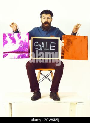 Bearded man, long beard. Brutal caucasian surprised hipster with moustache holding pink and orange shopping packages and sale inscription on blackboard, sitting on chair isolated on white background Stock Photo
