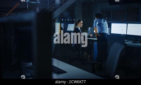 Confident Businessman Uses Desktop Computer, Her Male Manager Explains Specific Tasks, Account Handling and Strategy. Professional People Working Late Stock Photo