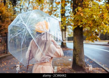 Fashionable woman in autumn clothes walks outdoors under transparent umbrella during rain. Fall season weather. Stock Photo