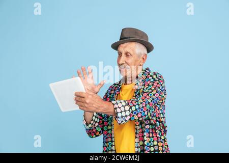 Videochat using tablet. Portrait of senior hipster man in stylish hat isolated on blue studio background. Tech and joyful elderly lifestyle concept. Trendy colors, forever youth. Copyspace for ad. Stock Photo