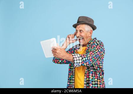 Videochat using tablet, shocked. Portrait of senior hipster man in stylish hat on blue studio background. Tech and joyful elderly lifestyle concept. Trendy colors, forever youth. Copyspace for ad. Stock Photo