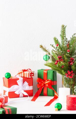 Red and green gift boxes and balls with candles , fir tree branches and hawthorn twigs in a vase on white wall background Stock Photo