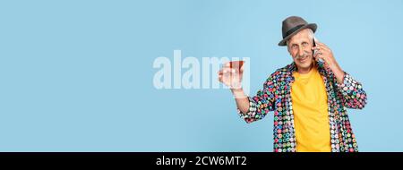 Drinks cocktail, talking phone. Portrait of senior hipster man in stylish hat on blue studio background. Tech and joyful elderly lifestyle concept. Trendy colors, forever youth. Copyspace, flyer Stock Photo