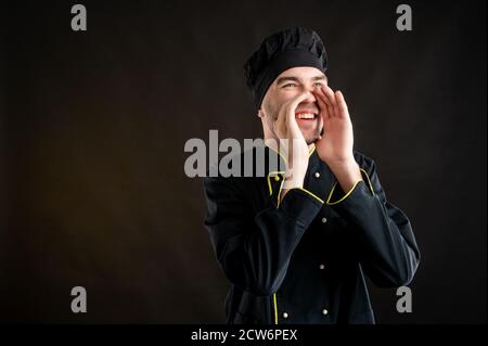 Portrait of young male dressed in a black chef suit shouting with his hands to his mouth posing on a brown background with copy space advertising area Stock Photo
