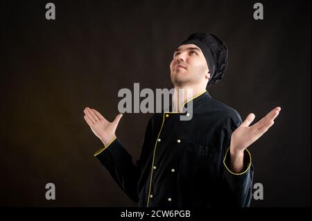 Portrait of young male dressed in a black chef suit with open arms looking up, side view posing on a brown background with copy space advertising area Stock Photo