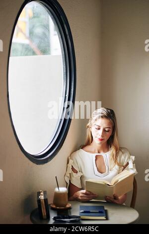 Charming smiling young woman sitting at table and reading interesting novel Stock Photo