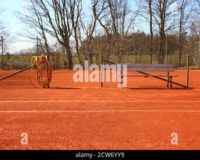 Detail of an empty red clay tennis court in the summer. There is a wooden bench and a yellow hose in the center. Stock Photo