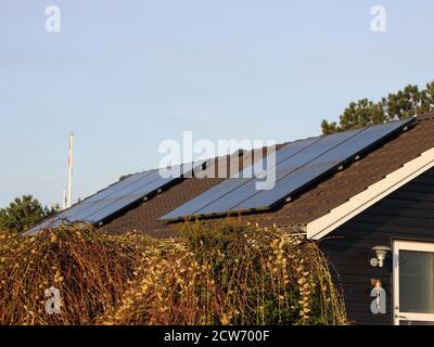 Two sets of solar cell panels on the roof of a black scandiniavian-style house. Bushes can be seen in the front and several flag poles are visible in Stock Photo