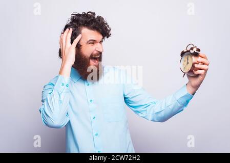 What is the hour, bearded man in casual looking afraid at alarm clock. Stock Photo