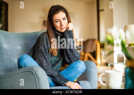 Portrait of smiling woman wearing turtleneck sweater and jeans while relaxing in the armchair at home. Stock Photo
