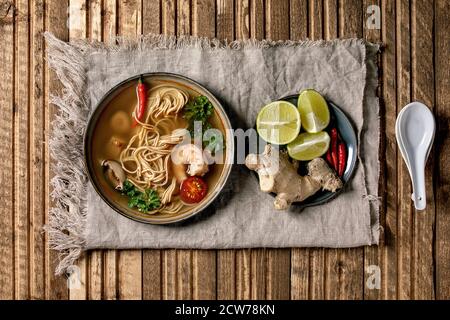 Traditional spicy Thai soup tom yum kung with shiitake mushrooms and prawns, ingredients in plate, spoon on linen cloth over wooden plank background. Stock Photo