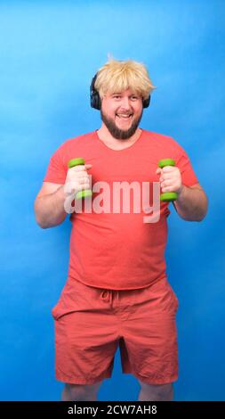 Funny chubby freak man in headphones and a wig in a pink t-shirt is engaged in green dumbbells and laughs on a blue background, vertical Stock Photo