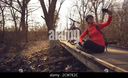 Happy caucasian woman cyclist makes a photo of herself a selfie on the phone while sitting on the bridge over a river in the forest in sunny weather Stock Photo
