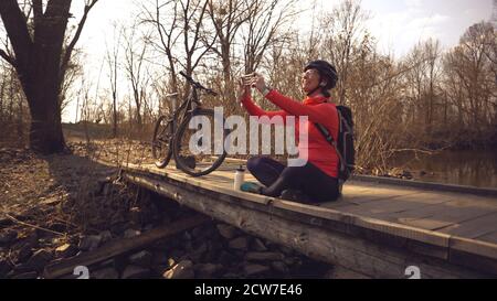 Happy caucasian woman cyclist makes a photo of herself a selfie on the phone while sitting on the bridge over a river in the forest in sunny weather Stock Photo