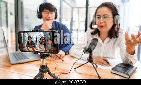 Asian Man and woman podcasters in headphones recording content with colleague talking to microphone and camera in broadcast studio together, communica Stock Photo