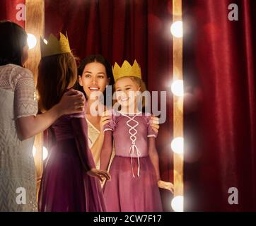 Cute little actress. Young mother and her daughter child girl in Princess costume on the background of theatrical scenes and mirrors. Stock Photo