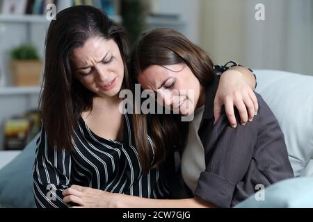 Two sad friends or sisters crying together on a couch in the living room at home