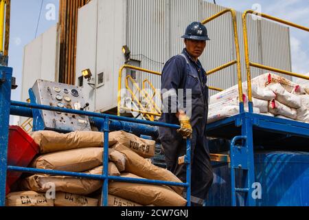 Oil deposit Zhaikmunai in desert. Worker in blue work wear and helmet with cement bags on mixer equipment. Oil rig on background. Blue sky with clouds Stock Photo