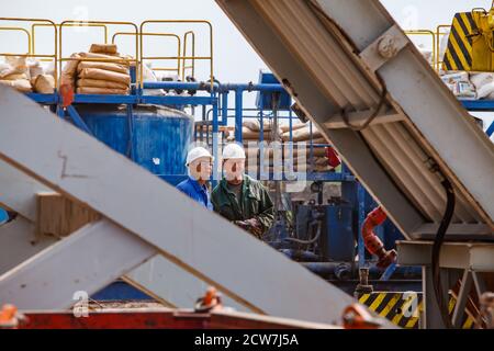 Oil deposit. Works oil drilling rig. Adult Serious Caucasian man and young Asian man in white hardhats stand on blue machine and cement paper bags bac Stock Photo