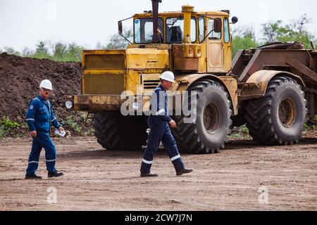 Two engineers or workers in blue work wear and white helmets on large yellow tractor background. Zhaik-Munai oil deposit, Kazakhstan. Stock Photo