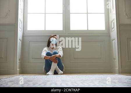 young asian woman in quarantine at home wearing facial mask sitting on floor legs crossed looking sad and depressed