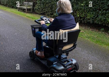 An elderly lady in a blue coat wearing a mask becasue of covid-19 enjoying the freedom of an electric mobility scooter on a quiet road. Stock Photo