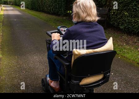 An elderly lady in a blue coat wearing a mask becasue of covid-19 enjoying the freedom of an electric mobility scooter on a quiet road. Stock Photo