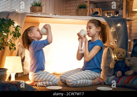 Two cute happy girls play at home. Funny lovely sisters are having fun in children room. Girls play tea-party and drink tea from cups. Stock Photo