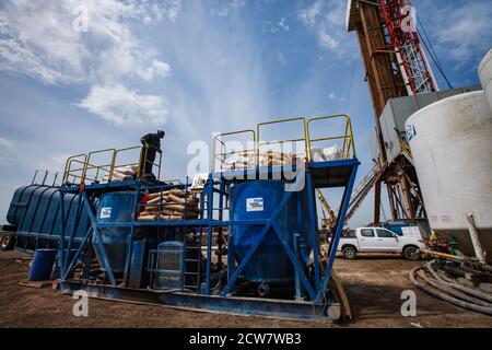 Oil deposit in desert. Worker in blue work wear and helmet with a cement in bags on mixer equipment. Oil rig on background. Blue sky with clouds. Pano Stock Photo