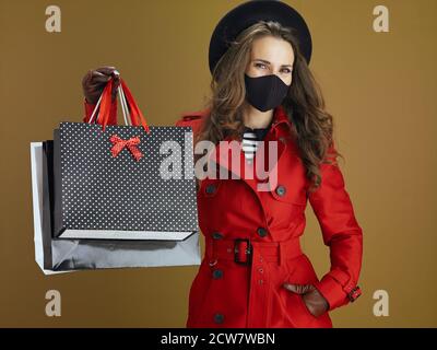 Hello autumn. Portrait of happy trendy middle aged housewife in red coat with black mask and paper shopping bags on beige background. Stock Photo