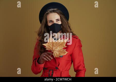 Hello autumn. pensive trendy middle aged housewife in red coat with black mask and yellow autumn maple leaf looking into the distance isolated on beig Stock Photo