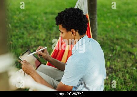 Side view of a student using digital tablet in park. Young freelancer drawing on digital tablet while sitting on a hammock. Stock Photo