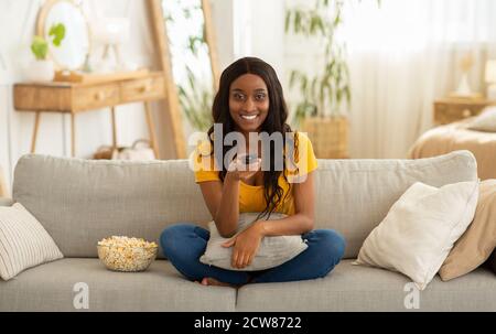 Home entertainments. Smiling black woman with remote control eating popcorn and watching TV on sofa in living room Stock Photo