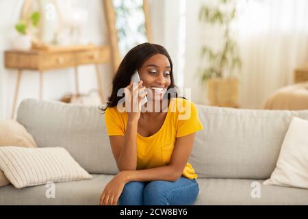 Attractive young black woman talking on mobile phone and smiling while sitting on sofa at home Stock Photo