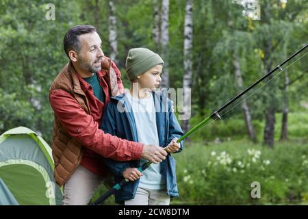 Side view portrait of mature father teaching son fishing while enjoying camping trip together by lake, copy space Stock Photo