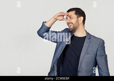 Young businessman man with a beard in a jacket, very happy and smiling, looking far away with hand above his head. Search concept. Portrait of a man on a gray background. Stock Photo
