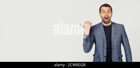 Young businessman man with a beard in a jacket, surprised, pointing finger to the side, with open mouth amazed expression. Portrait of a man on a gray background. Stock Photo