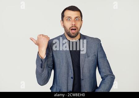 Young businessman man with a beard in a jacket, surprised, pointing finger to the side, with open mouth amazed expression. Portrait of a man on a gray background. Stock Photo