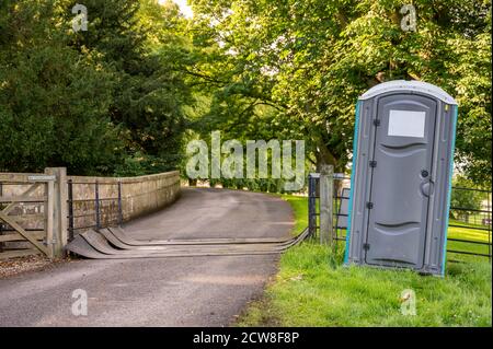 A grey plastic portable toilet next to a covered cattle grid on a public footpath in a field at an outdoor event