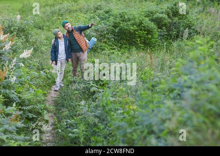 Portrait of father and son hiking together with man talking to boy and pointing away while walking on wild path in forest, copy space Stock Photo