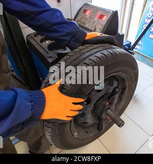 Mechanic working with wheel balancing machine at tire service, closeup. Mechanic wearing orange gloves and blue outfit. Stock Photo